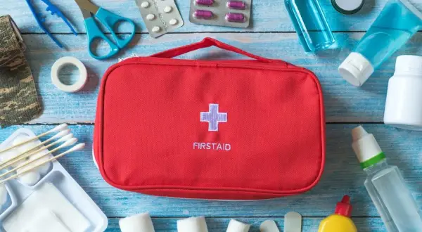 Czerwona torba z napisem FIRSTAID i krzyżem, obok niej wyposażenie apteczki samochodowej: plastry, bandaże, nożyczki, pęseta, leki, płyny.