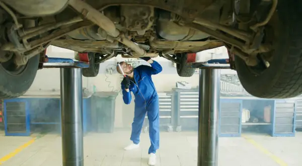 Mechanik w niebieskim kombinezonie bada podwozie samochodu na podnośniku. W powietrzu unosi się lekka mgiełka, przypominająca zapach benzyny w samochodzie.