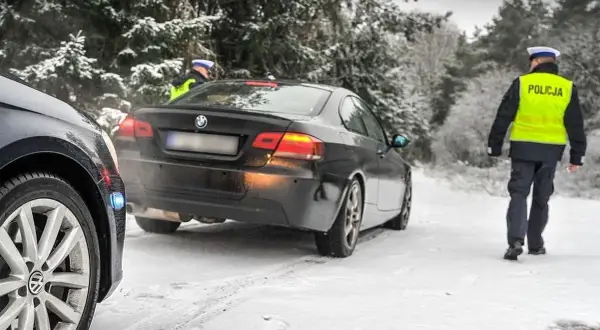 Policjant w kamizelce odblaskowej idzie w stronę czarnego BMW na zaśnieżonej drodze. Kierowca pewnie dostanie mandat za driftowanie.