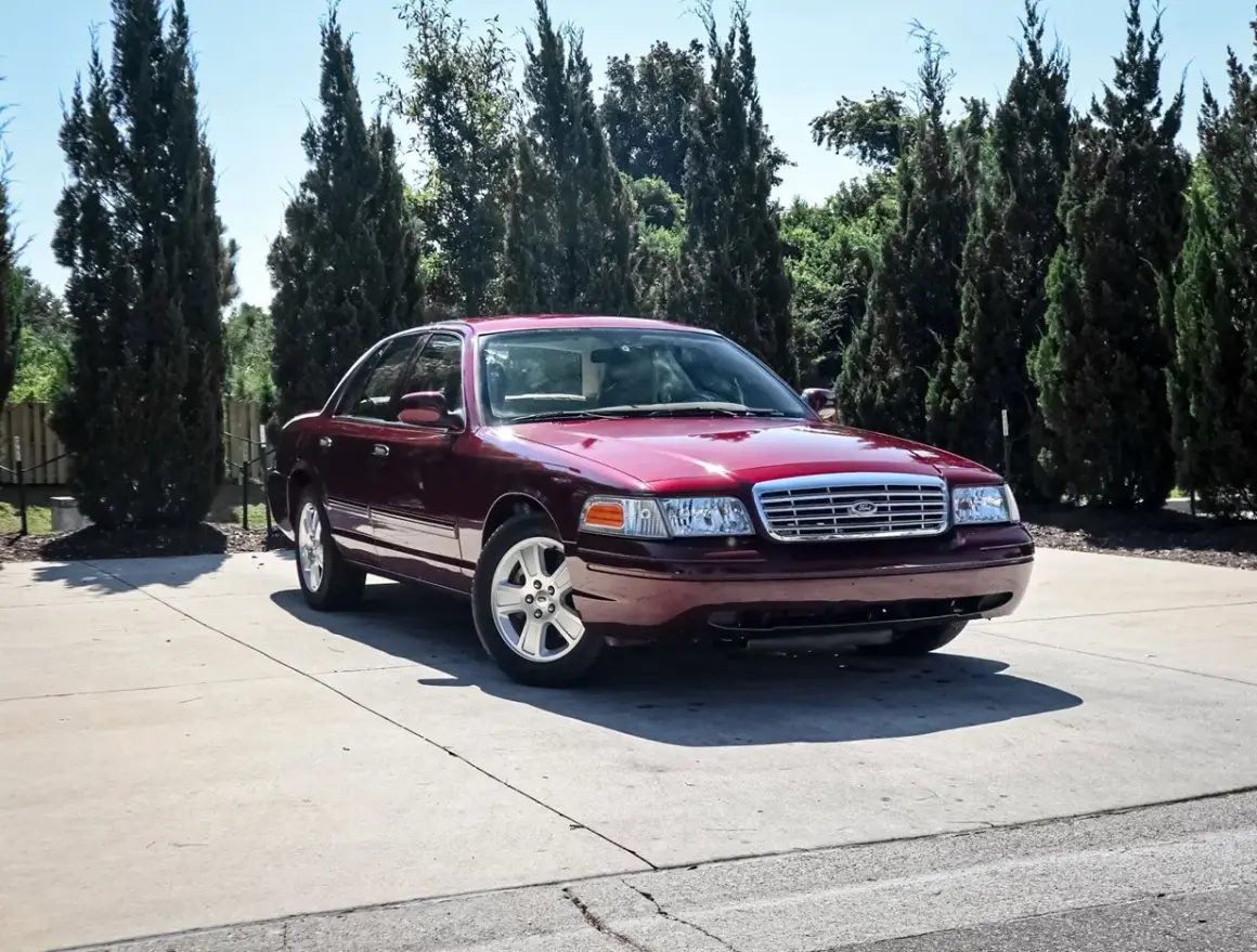 Ciemnoczerwony Ford Crown Victoria, klasyk z USA, zaparkowany na podjeździe wśród wysokich drzew.