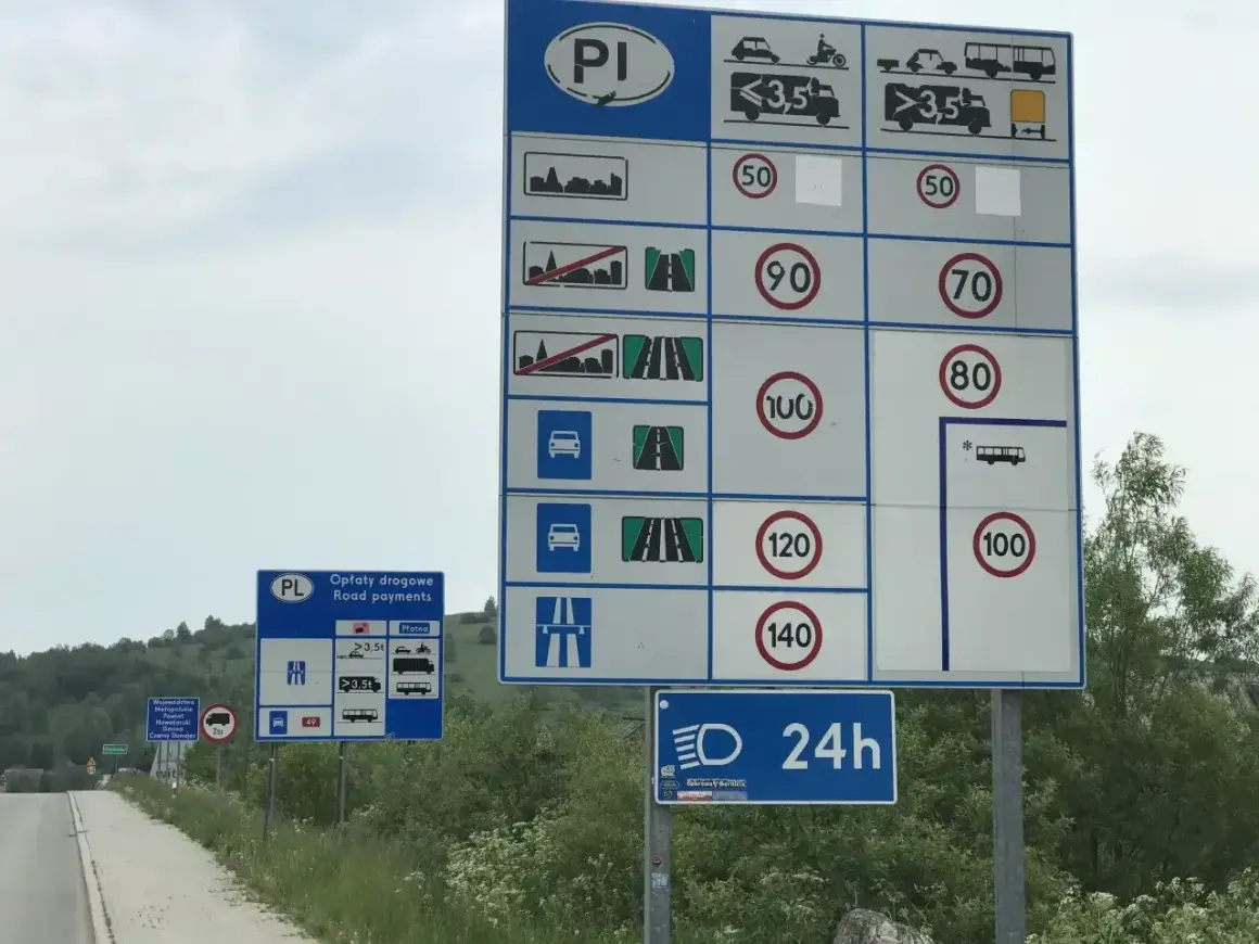 Znaki drogowe informują o ograniczeniu w terenie zabudowanym, opłatach drogowych i limitach prędkości dla różnych pojazdów.