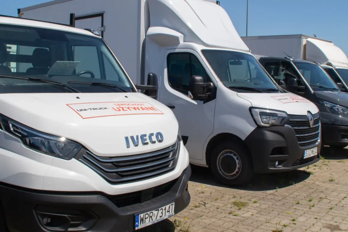 Rząd białych i ciemnych samochodów osobowo dostawczych zaparkowanych na zewnątrz. Na masce białego Iveco widnieje napis "Samochody Używane".