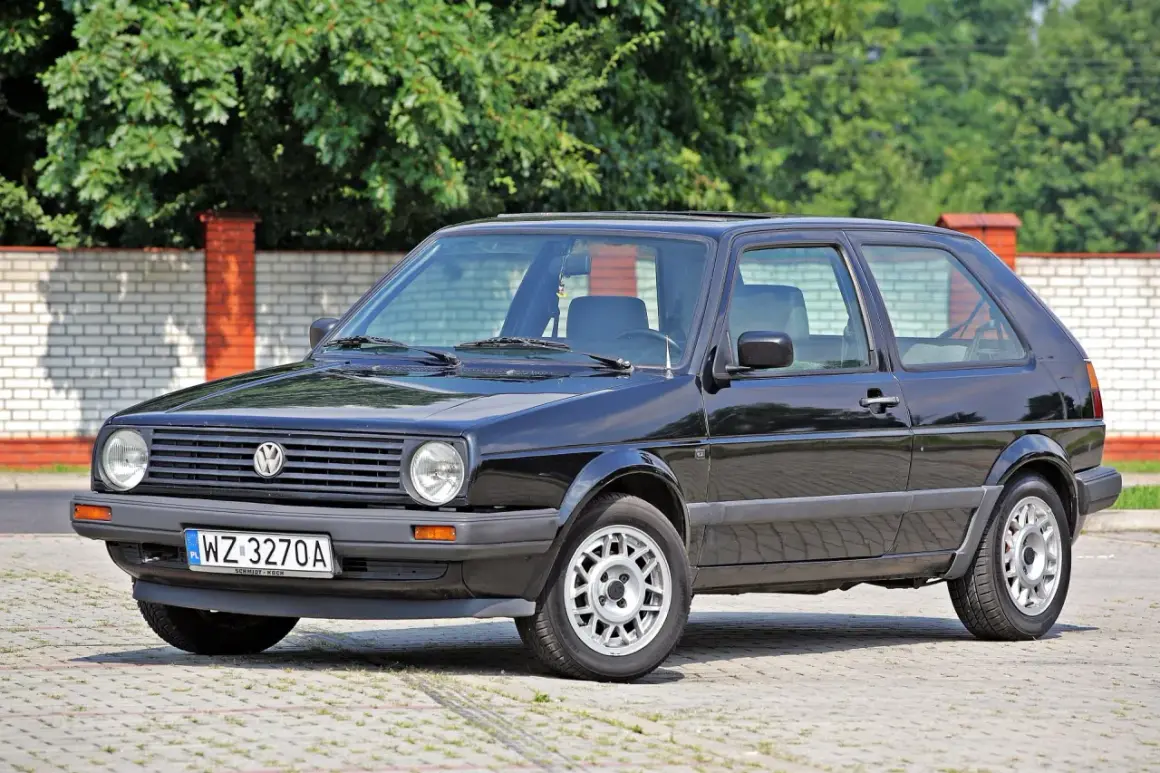 Czarny VW Golf 2, klasyk z lat 80., lśni w słońcu na tle ceglanego muru i zieleni.