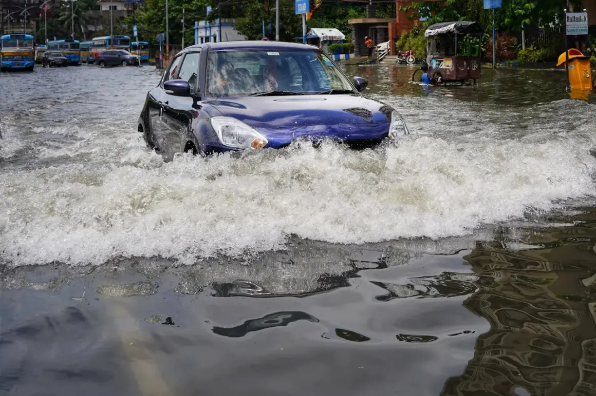 Niebieskie auto walczy z falą wody na zalanej ulicy. W tle widać inne auta po powodzi i autobusy.