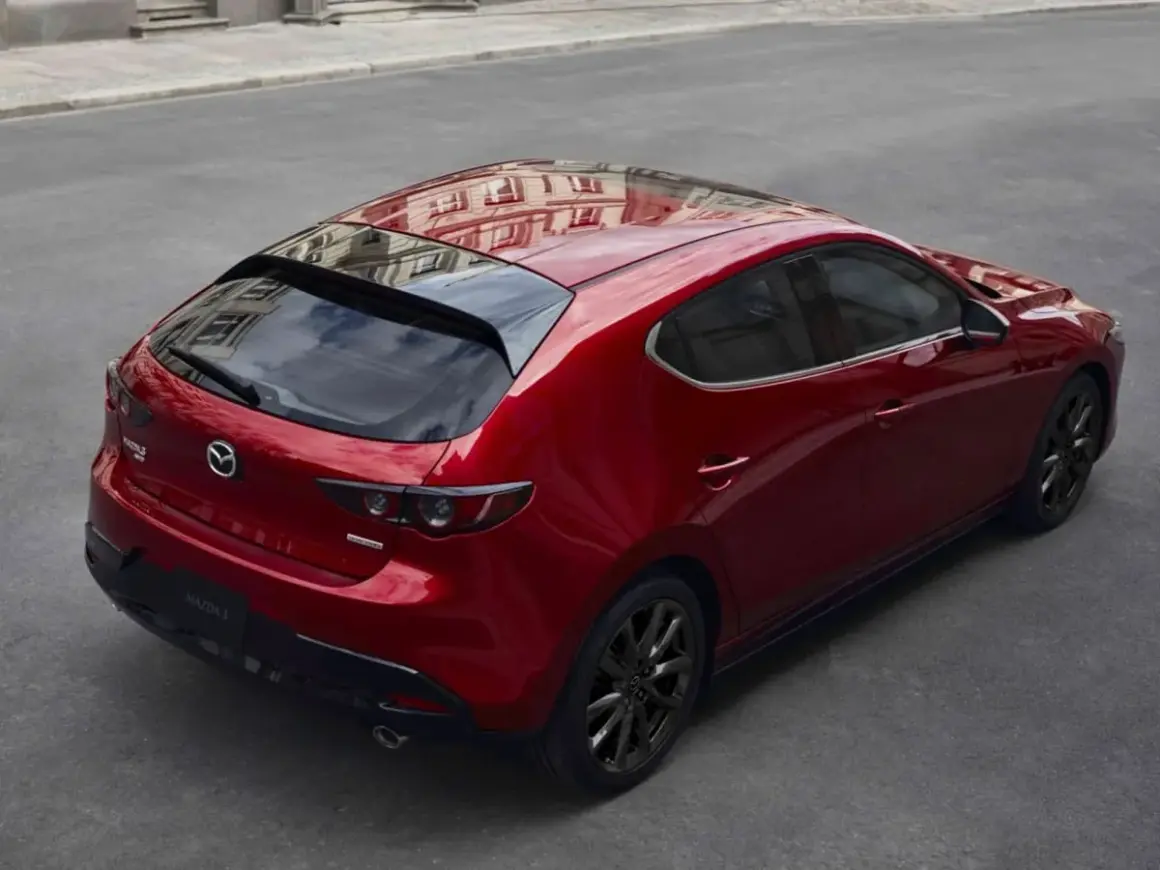 Czerwony Mazda 3 hatchback z czarnymi felgami, odbijający budynki. Elegancki, sportowy design, który odróżnia go od sedana.