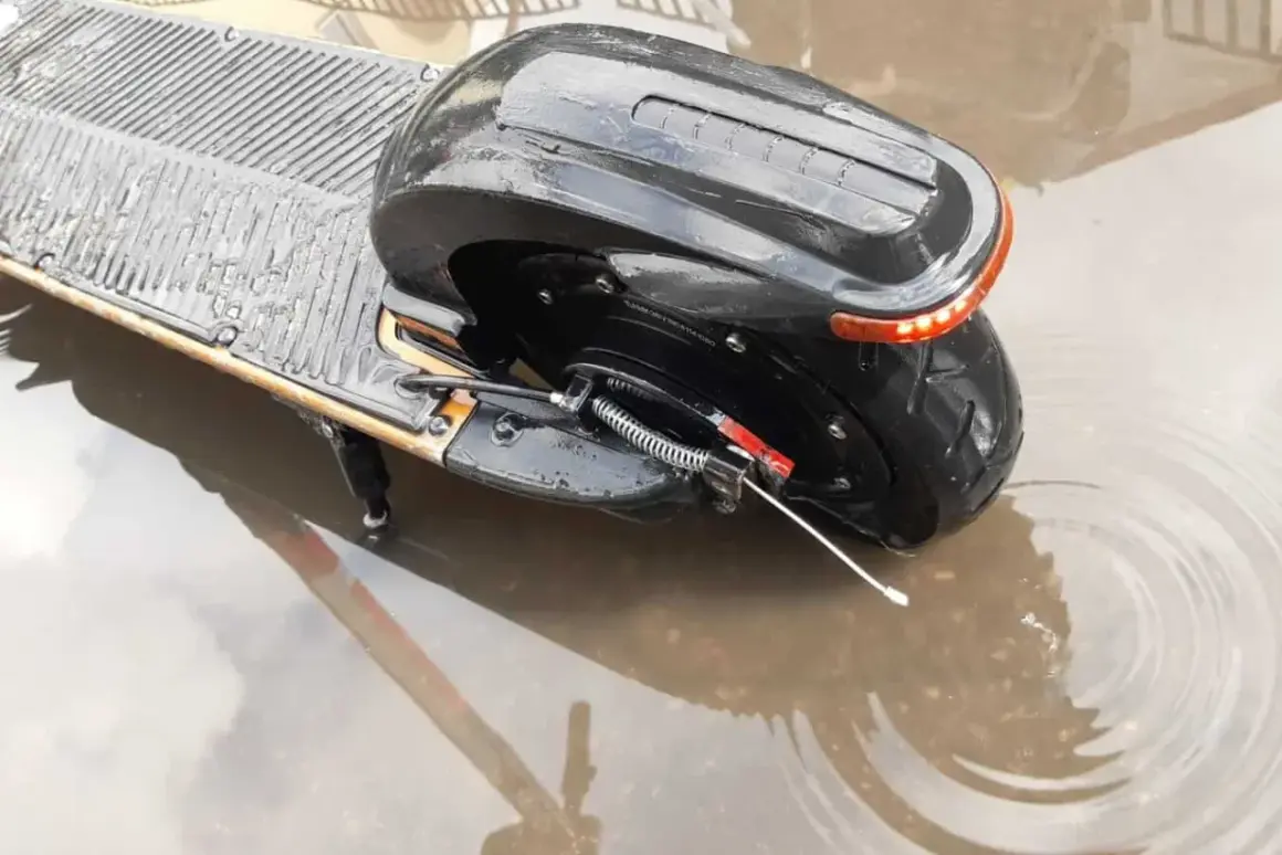 Czarna hulajnoga elektryczna stoi w kałuży. Czy hulajnoga elektryczna można jeździć w deszczu? Woda sięga do tylnego koła i części podestu.
