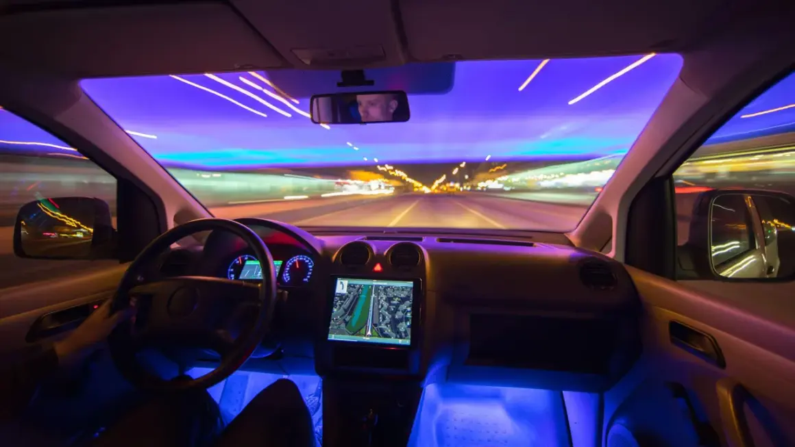 Wnętrze samochodu nocą, z widokiem na drogę i światła miasta. Kierowca obserwuje nawigację, przygotowując się na przyszłość autonomicznych pojazdów.