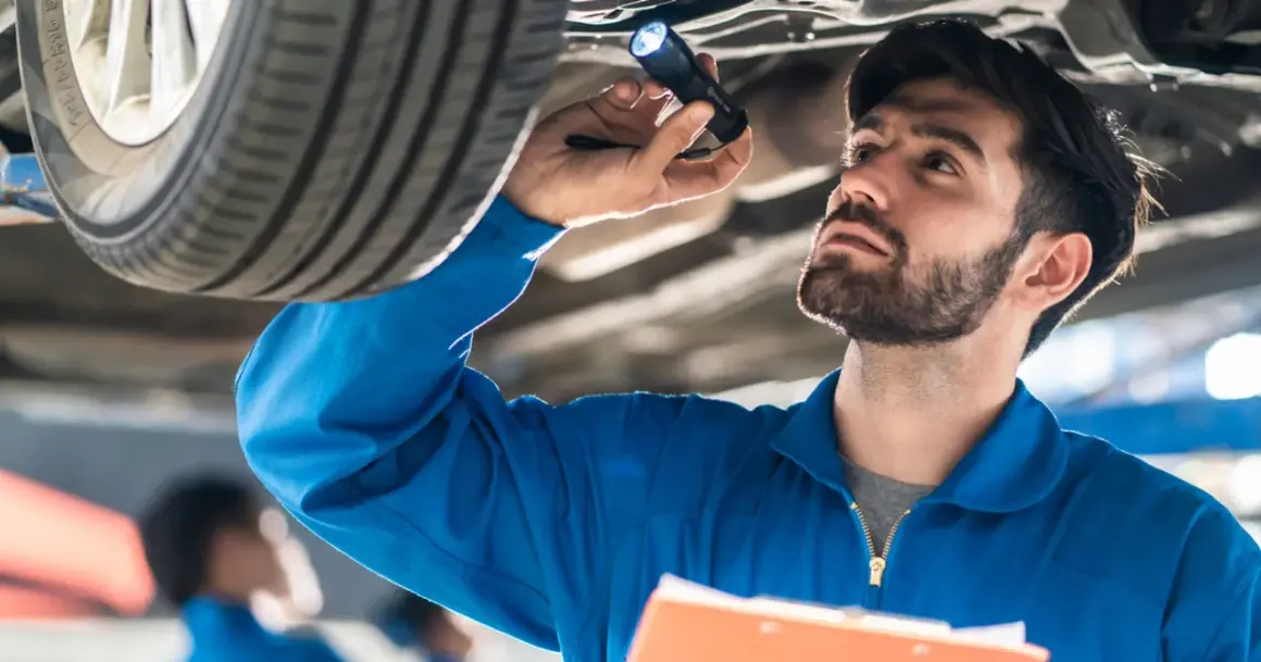Mechanik w niebieskim kombinezonie oświetla latarką podwozie samochodu, wykonując **serwisowanie samochodu**.