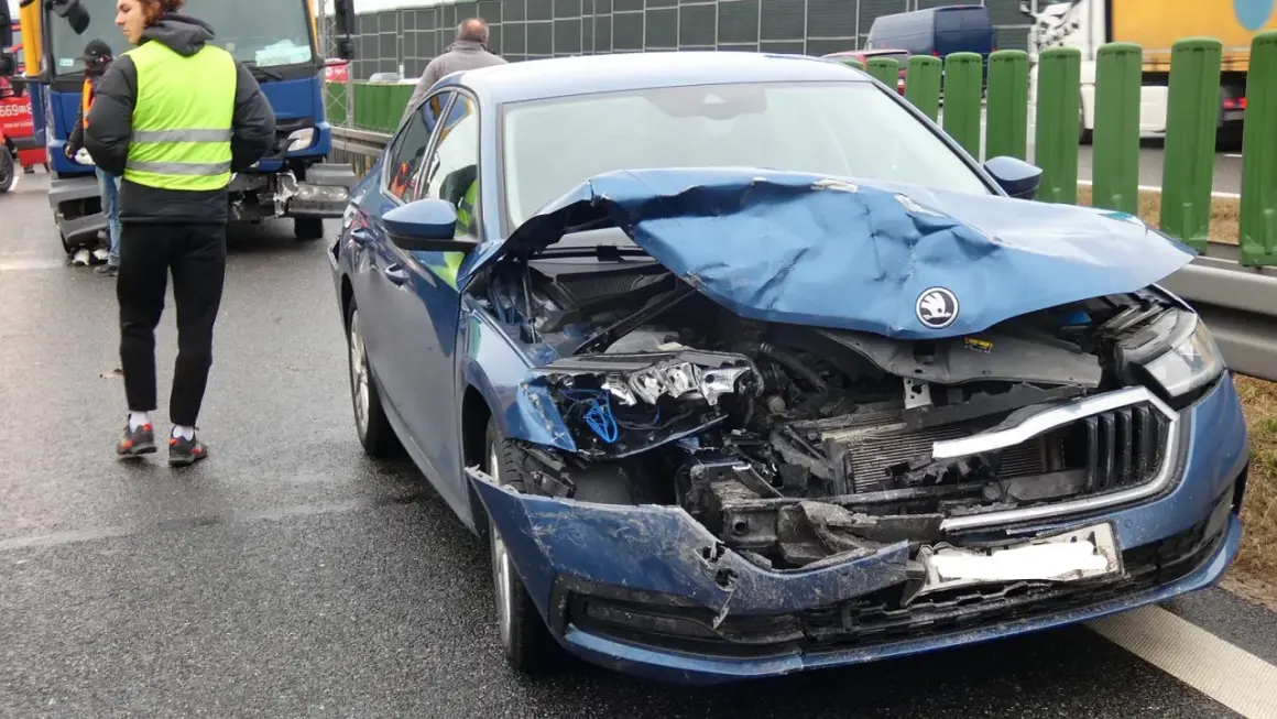Widok na zniszczony przód niebieskiego samochodu po wypadku samochodowym. Na drodze widać osoby w kamizelkach odblaskowych.