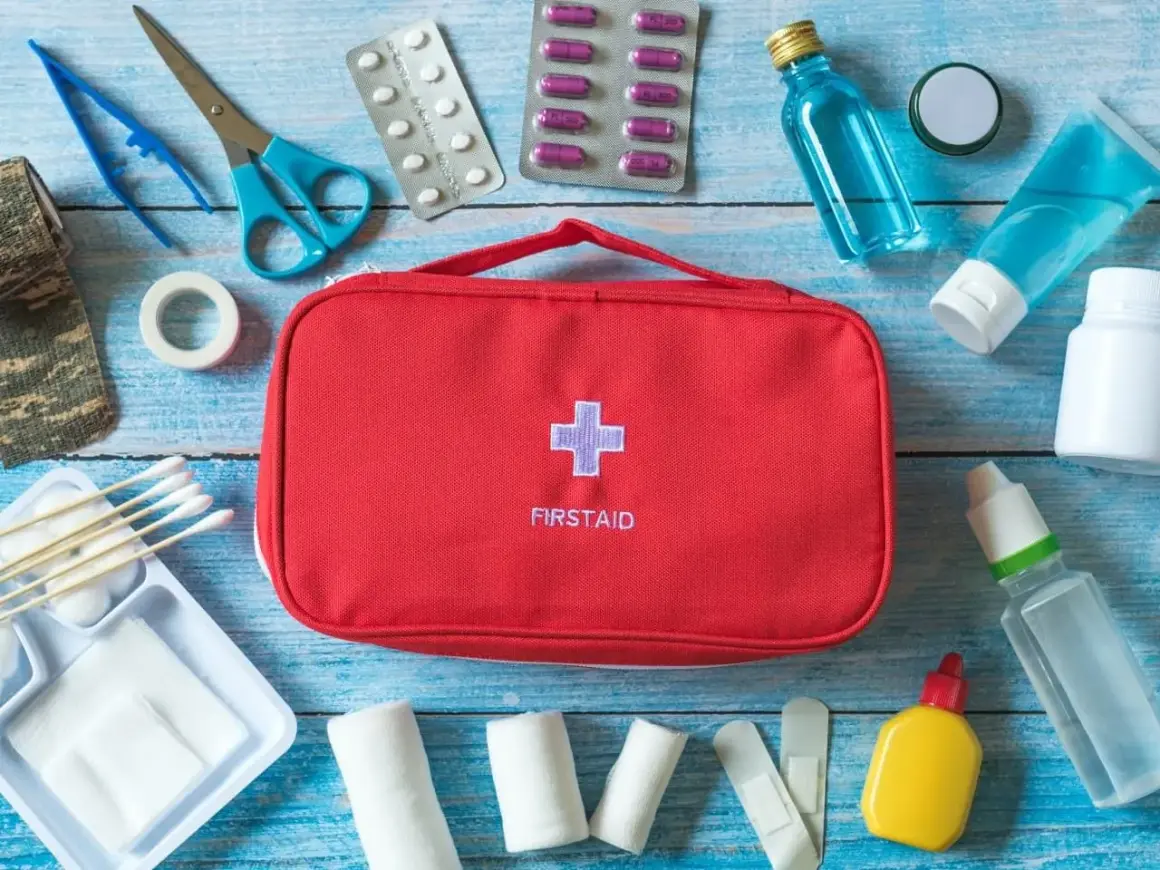 Czerwona torba z napisem FIRSTAID i krzyżem, obok niej wyposażenie apteczki samochodowej: plastry, bandaże, nożyczki, pęseta, leki, płyny.