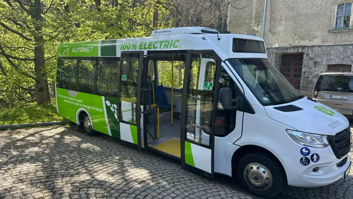 Nowy, zielono-biały bus elektryczny "Automety" z napisem "100% ELECTRIC" stoi na bruku.