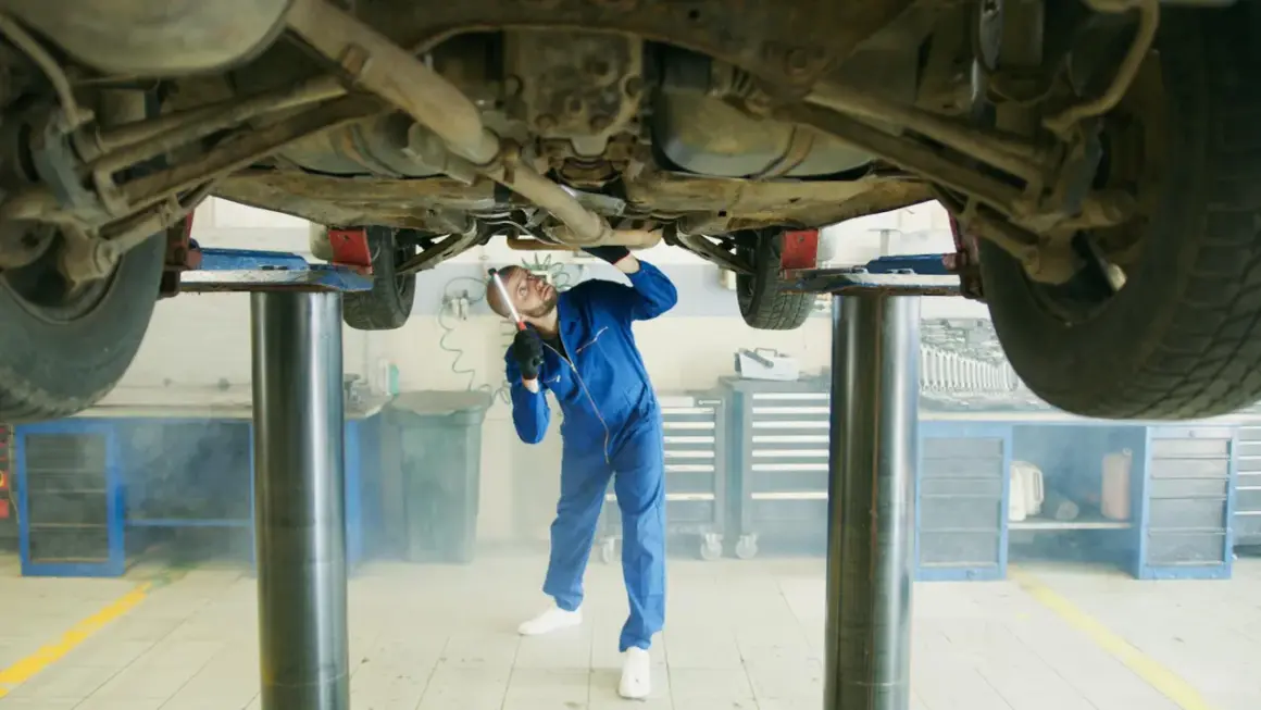 Mechanik w niebieskim kombinezonie bada podwozie samochodu na podnośniku. W powietrzu unosi się lekka mgiełka, przypominająca zapach benzyny w samochodzie.