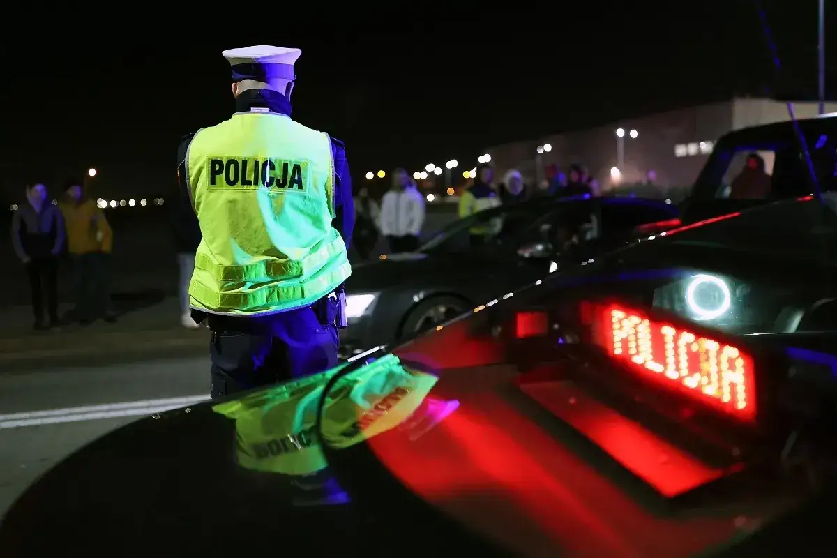 Policjant w kamizelce odblaskowej kontroluje ruch. Na dachu radiowozu świeci napis 