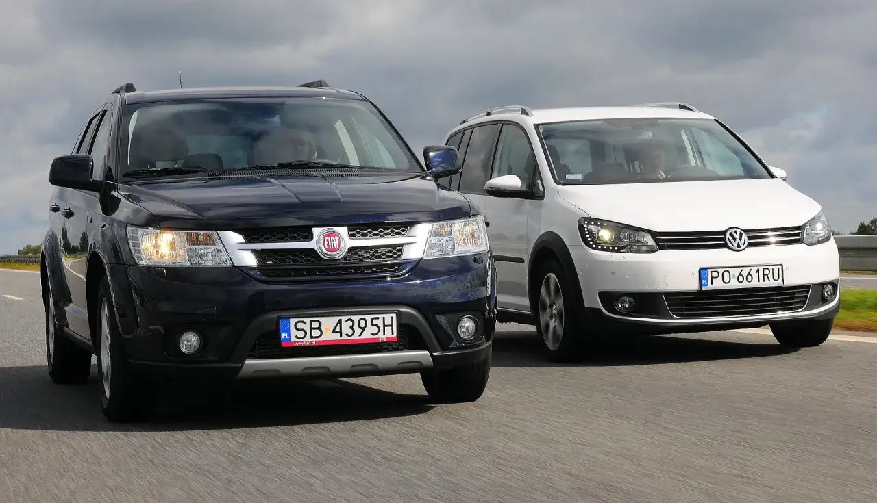 Ciemny Fiat i biały Volkswagen, oba to 6 osobowe auta, jadą obok siebie po drodze.