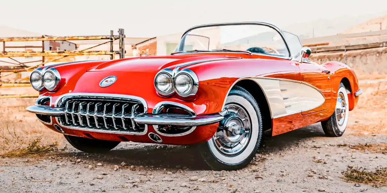 Klasyczny czerwony kabriolet Corvette, ikona amerykańskiej marki samochodów, lśni chromem na tle wiejskiego krajobrazu.