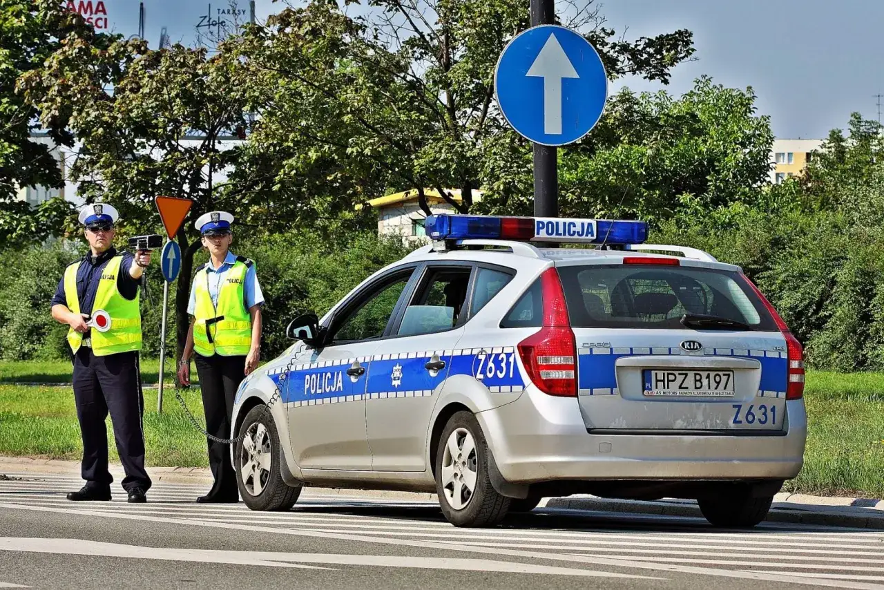 Dwóch policjantów w kamizelkach odblaskowych i radiowóz z włączonymi sygnałami świetlnymi. Czy radiowóz musi być widoczny? Tak, dla bezpieczeństwa.