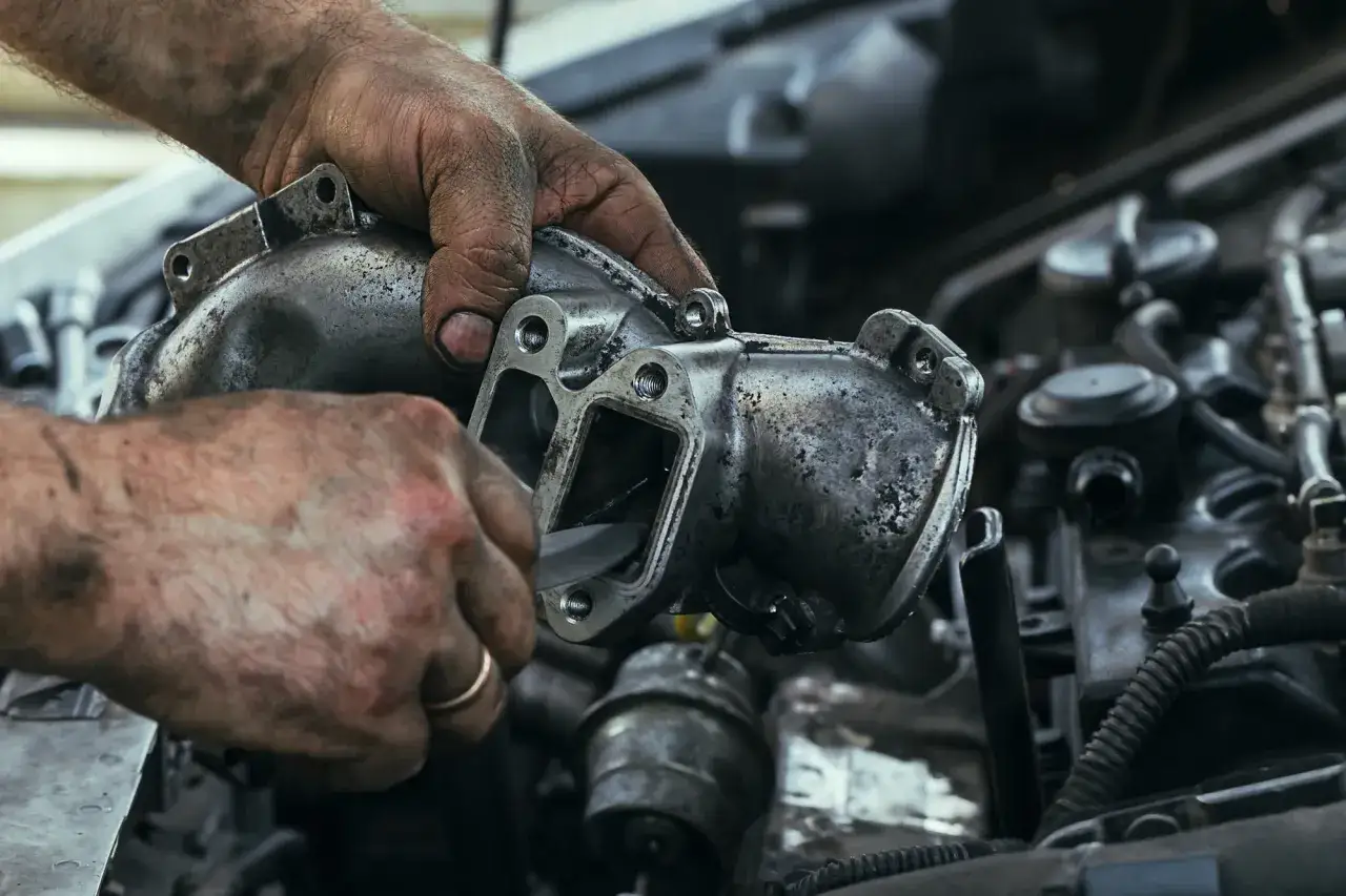 Brudne ręce mechanika czyszczą zapieczony zawór EGR. Widać narzędzie wkładane do wnętrza elementu.