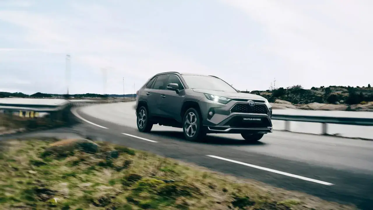 Szary SUV Toyota RAV4 Plug-in Hybrid, hybryda plug-in z największym zasięgiem, jedzie po krętej drodze nad wodą.