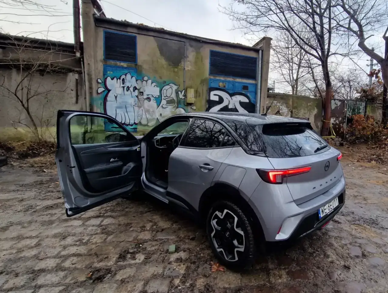 Srebrny Opel Mokka z otwartymi drzwiami, gotowy do darowizny samochodu po leasingu, na tle zniszczonego budynku z graffiti.