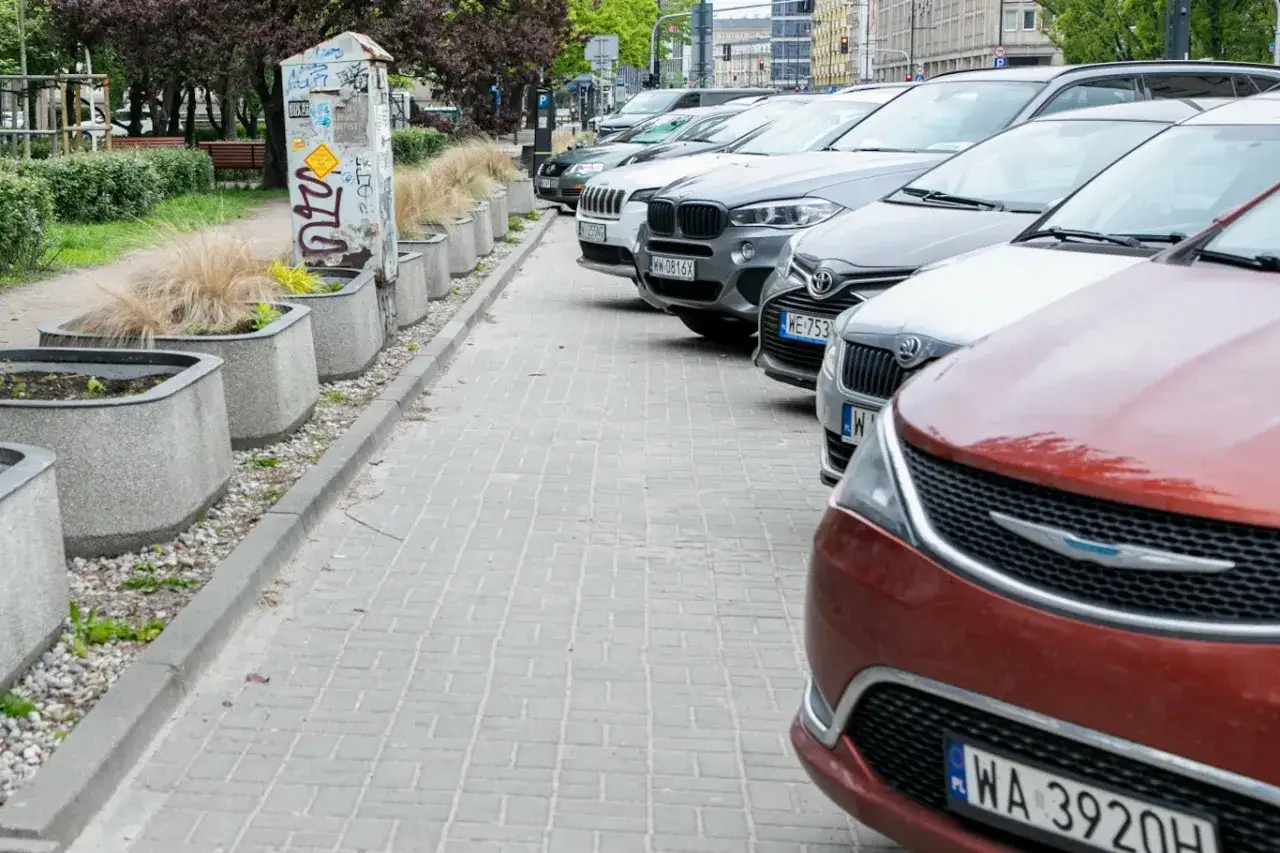 Rząd samochod&oacute;w zaparkowanych na chodniku, obok donic z trawą. Czy można parkować na chodniku? To pytanie nasuwa się samo.