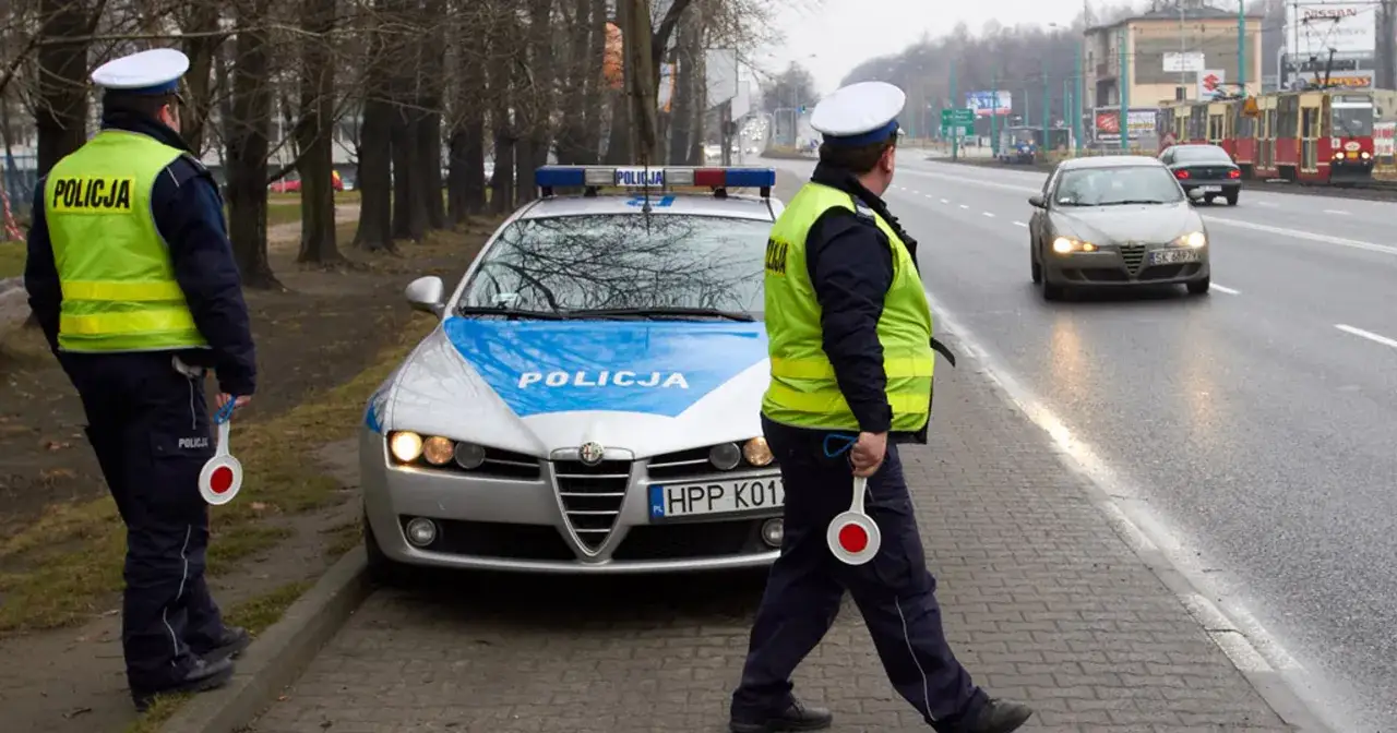 Policjanci zatrzymują kierowcę z powodu przekroczenia prędkości. Na drodze stoi radiowóz, a jeden z funkcjonariuszy trzyma lizak do zatrzymywania pojazdów.