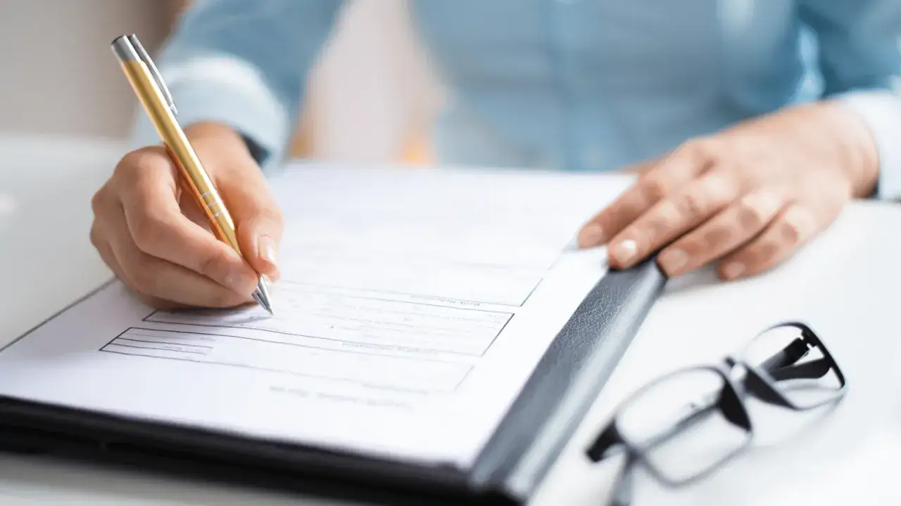 Dłoń z długopisem wypełnia formularz, sugerując prosty leasing. Obok leżą okulary.