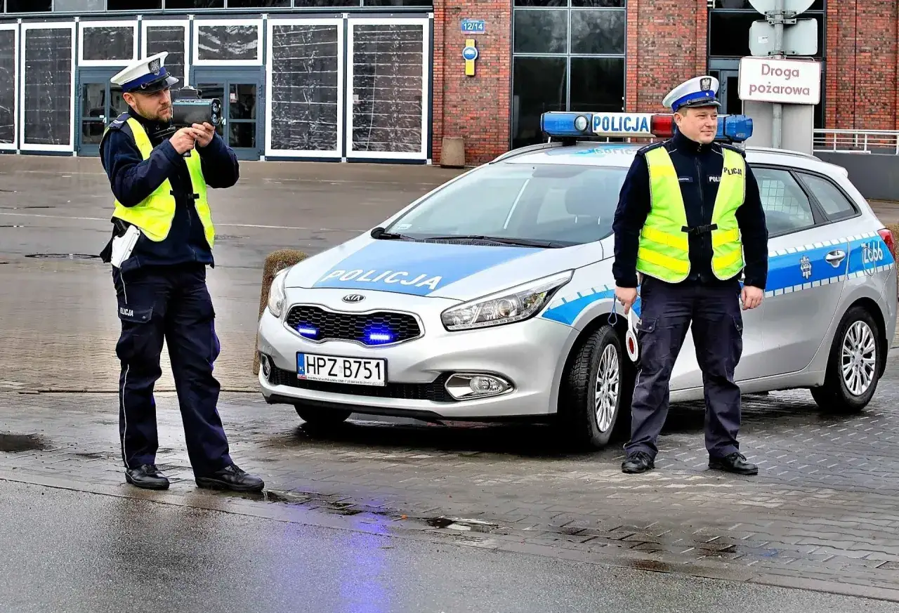 Policjant z radarem mierzy prędkość z pewnej odległości. Obok radiowóz z włączonymi niebieskimi światłami.
