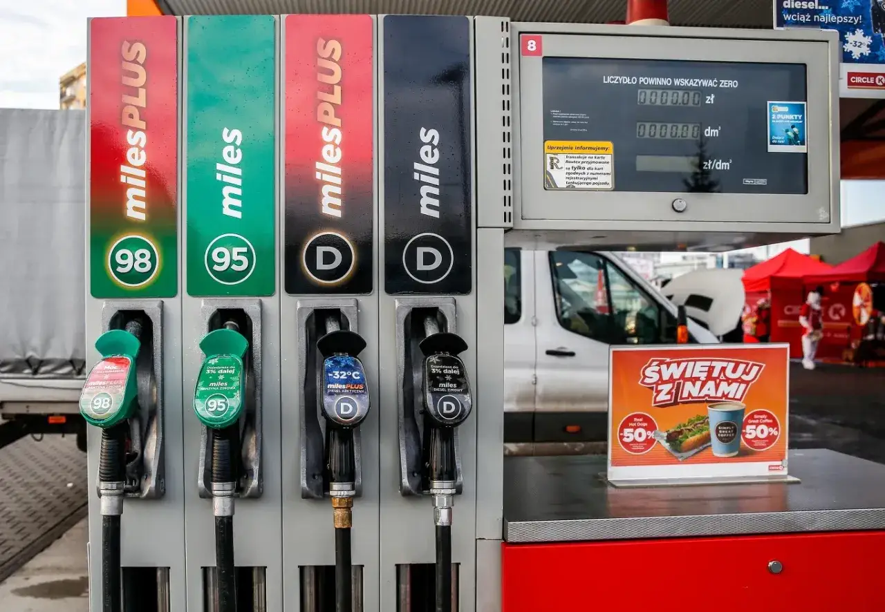 Stacja paliw z dystrybutorami benzyny 98, 95 i diesel. Widać pistolety do tankowania, ekran z cenami oraz reklamę hot doga i kawy.