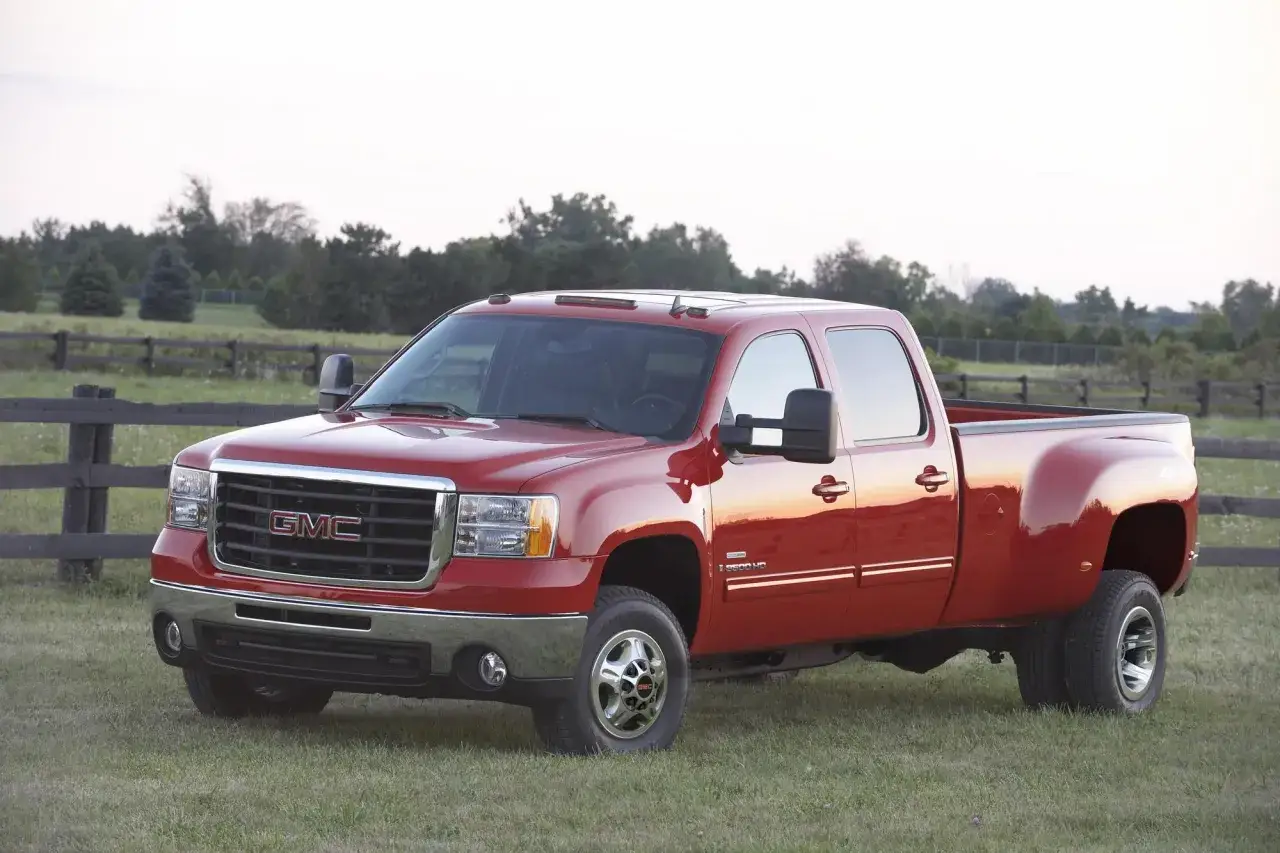Czerwony pickup amerykański GMC Sierra HD na trawiastym polu, z drewnianym płotem w tle.