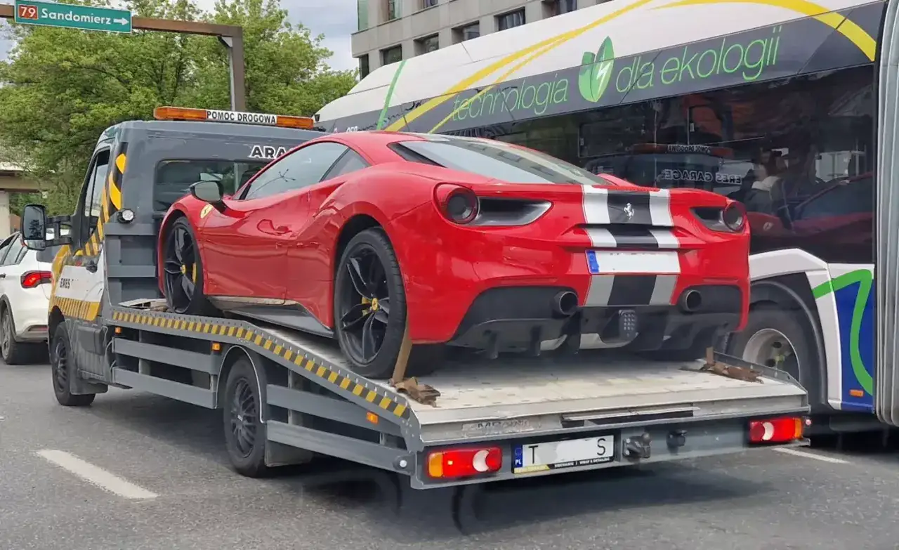 Czerwone Ferrari na lawecie, przygotowywane do sprzedaży samochodu po leasingu. Obok autobus z napisem