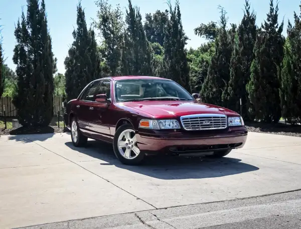 Ciemnoczerwony Ford Crown Victoria, klasyk z USA, zaparkowany na podjeździe wśród wysokich drzew.