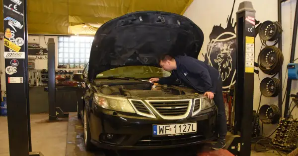 Mechanik sprawdza silnik czarnego Saaba. Na podnośniku warsztatowym, w tle widać narzędzia i części samochodowe.