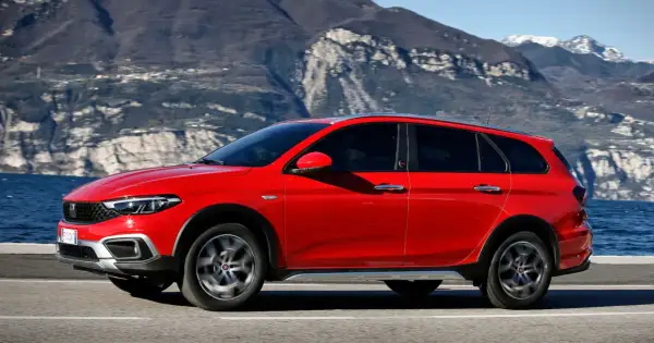 Czerwony Fiat Tipo Cross, wszechstronny samochód z segmentu kombi, przemierza malowniczą drogę nad jeziorem, z górami w tle.