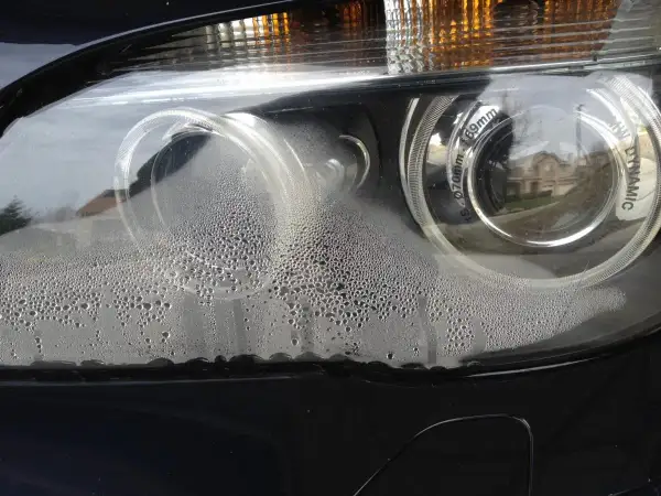 Zaparowany reflektor od środka, z licznymi kropelkami wody na kloszu. Widać odbicie domu i drzew.