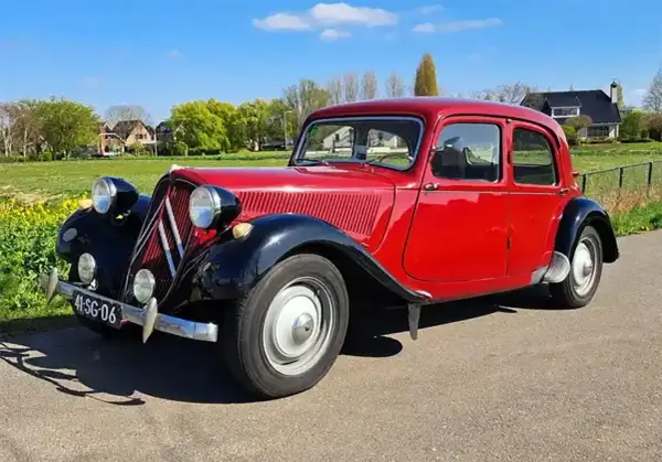 Klasyczny, czerwony Citroen Traction Avant, jeden z kultowych modele stare, zaparkowany na tle zielonej łąki i domów.
