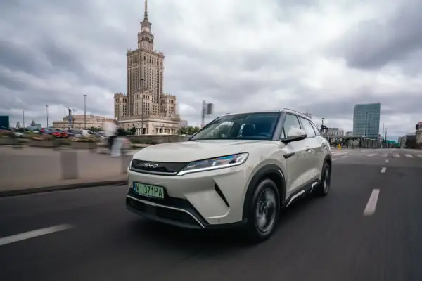 Biały SUV z logo chińskich samochodów BYD jedzie ulicą w Warszawie, z Pałacem Kultury w tle.