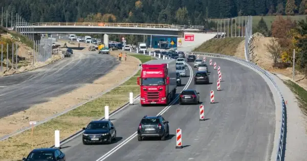 Budowa zakopianki w otoczeniu lasu. Ruch samochodowy, w tym czerwona ciężarówka, odbywa się po nowej drodze z mostem w tle.