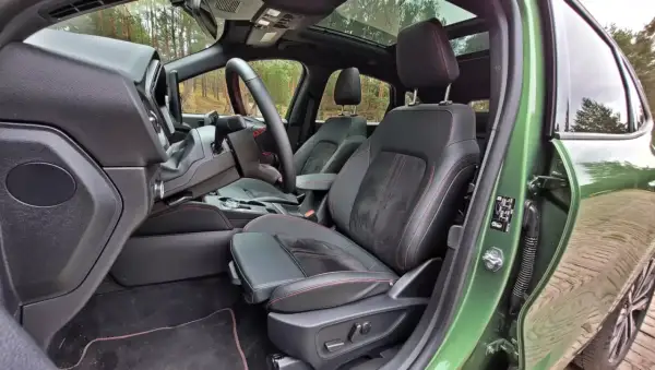 Wnętrze Ford Kuga z czarną tapicerką i czerwonymi przeszyciami. Widok na fotel kierowcy, deskę rozdzielczą i fragment dachu panoramicznego.