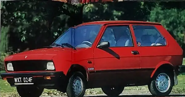 Czerwony yugo samochód z czarną atrapą chłodnicy i przyciemnianymi szybami, zaparkowany na tle zieleni.