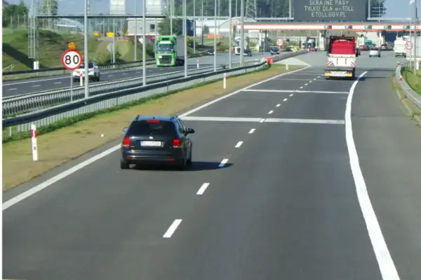 Czarny samochód jedzie autostradą, zbliżając się do bramek e-TOLL. Znak ograniczenia prędkości do 80 km/h przypomina o zachowaniu ostrożności.