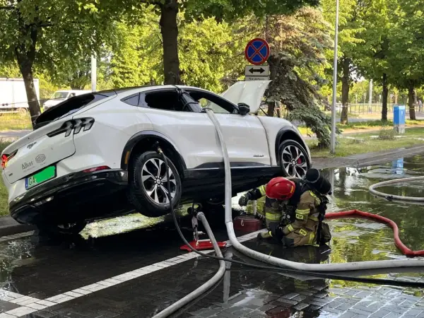 Strażak w akcji, gaszenie samochodu elektrycznego. Woda i węże strażackie wokół białego SUV-a.