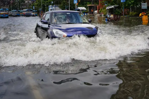 Niebieskie auto walczy z falą wody na zalanej ulicy. W tle widać inne auta po powodzi i autobusy.