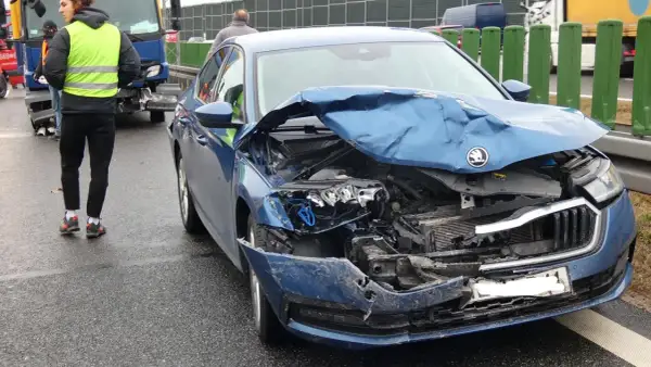 Widok na zniszczony przód niebieskiego samochodu po wypadku samochodowym. Na drodze widać osoby w kamizelkach odblaskowych.