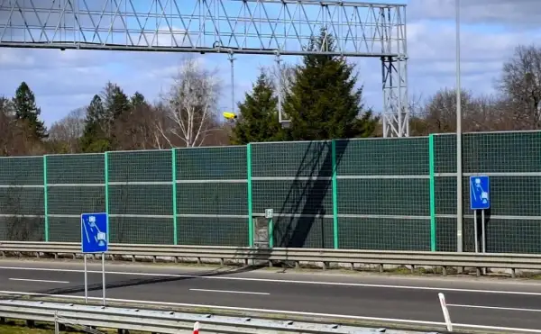 Znak informuje o odcinkowym pomiarze prędkości S1. Za zieloną barierą dźwiękochłonną widać drzewa i niebo.